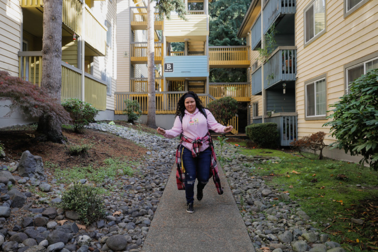 Washington homelessness and foster care: Young woman with long, dark, curly hair in pink sweatshirt and jeans, skips down an ivy-lined cement walkway between two tall apartment buildings.