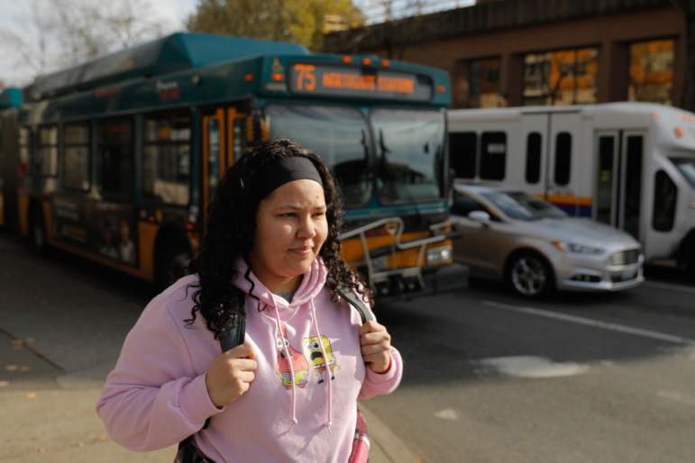 Washington homelessness and foster care: Young woman with long, dark, curly hair in pink sweatshirt and daypack walks along city sidewalk with two city busses in the background.