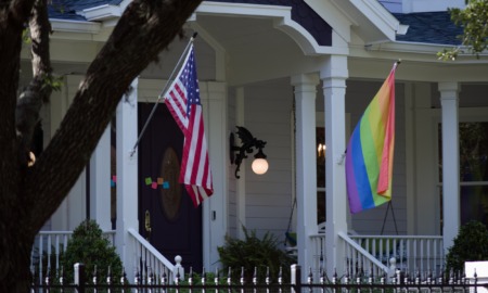 LGBTQ+ safe homes: Traditional style architecture home closeup of white front porch with black door, stairs and two pillars — one with American flag the other with a rainbow flag.