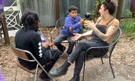 Youth podcaster: Three people outside in lawn chairs — one black-haired preteen in all black, and one preteen with dark hair in a marine blue sweatshirt holding microphone to dark-haired adult in black overalls gesturing with hands while speaking