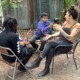Youth podcaster: Three people outside in lawn chairs — one black-haired preteen in all black, and one preteen with dark hair in a marine blue sweatshirt holding microphone to dark-haired adult in black overalls gesturing with hands while speaking