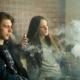 Vaping Teens & Law: Teen girl and boy sit leaning against a red brick wall vaping , heads surrounded by smoke.