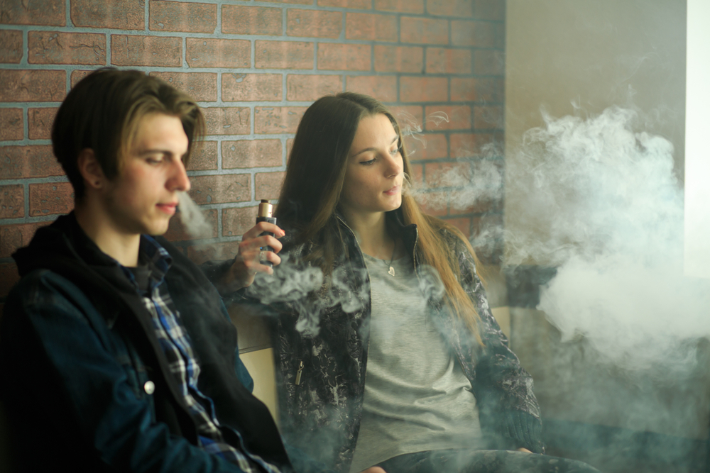 Vaping Teens & Law: Teen girl and boy sit leaning against a red brick wall vaping , heads surrounded by smoke.