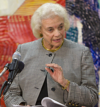 iCivics curriculum: Justice Sandra Day O'Connor, an older, woman with bobbed gray hair. wearing black and white checked jacket, stands at podium with microphone gesturing with her left hand as she speaks.