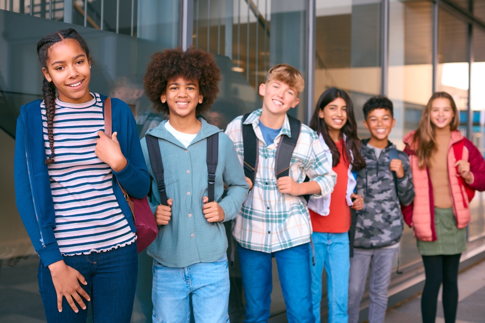 Happiness psychology: Line of happy teens stand with backpacks and book in front of a wall with floor to ceiling windows