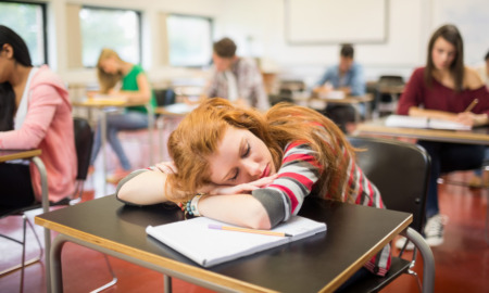 Syudent sleep depravation: Reen redhead girl sleeps with hed on arms sitting at classroom vesk surrounded ny other students at desks
