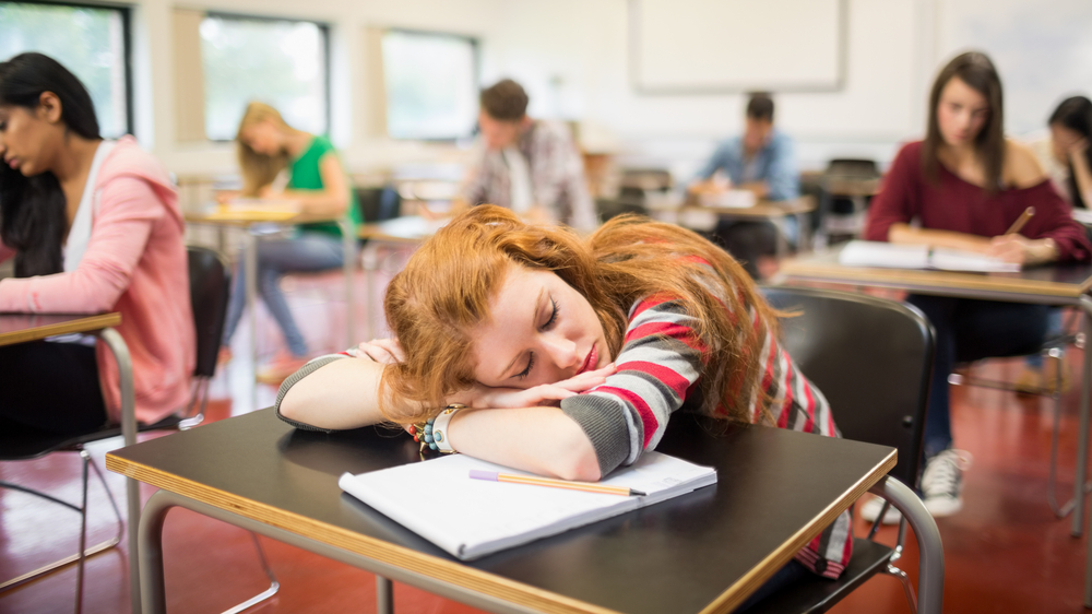 Syudent sleep depravation: Reen redhead girl sleeps with hed on arms sitting at classroom vesk surrounded ny other students at desks