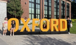 Affirmative Action: Several adults sit on or stand next to a very large, dark yellow,3D letter sign spelling "OXFORD" standing on the lawn in front of a multi-story red brick building o a sunny day.