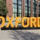 Affirmative Action: Several adults sit on or stand next to a very large, dark yellow,3D letter sign spelling "OXFORD" standing on the lawn in front of a multi-story red brick building o a sunny day.