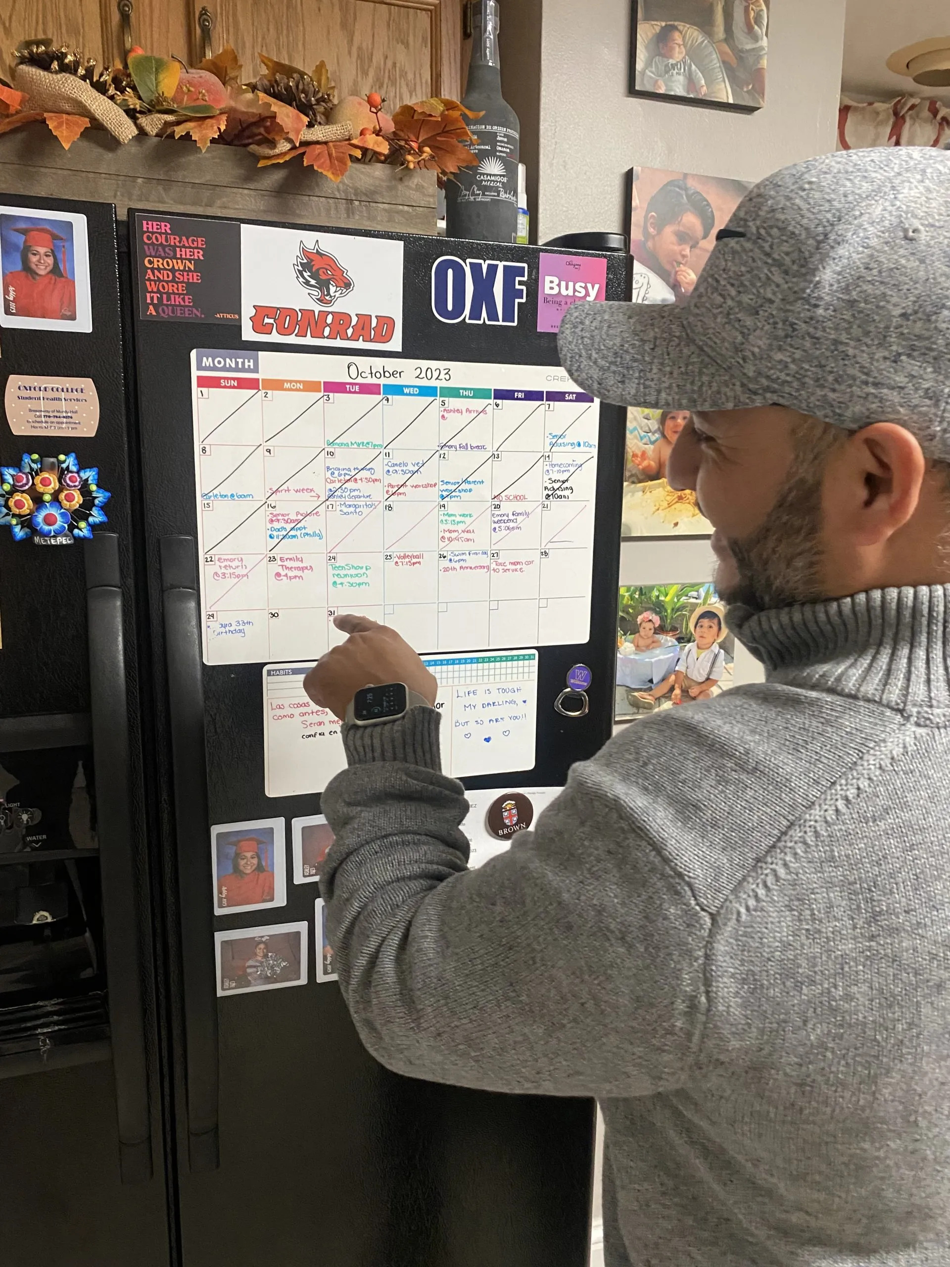 Closeup of man in profile with baseball cap and gray sweatshirt standing in front of black refrigerator pointing to a large family calendar surrounded by photos of family and stickers on the refrigerator