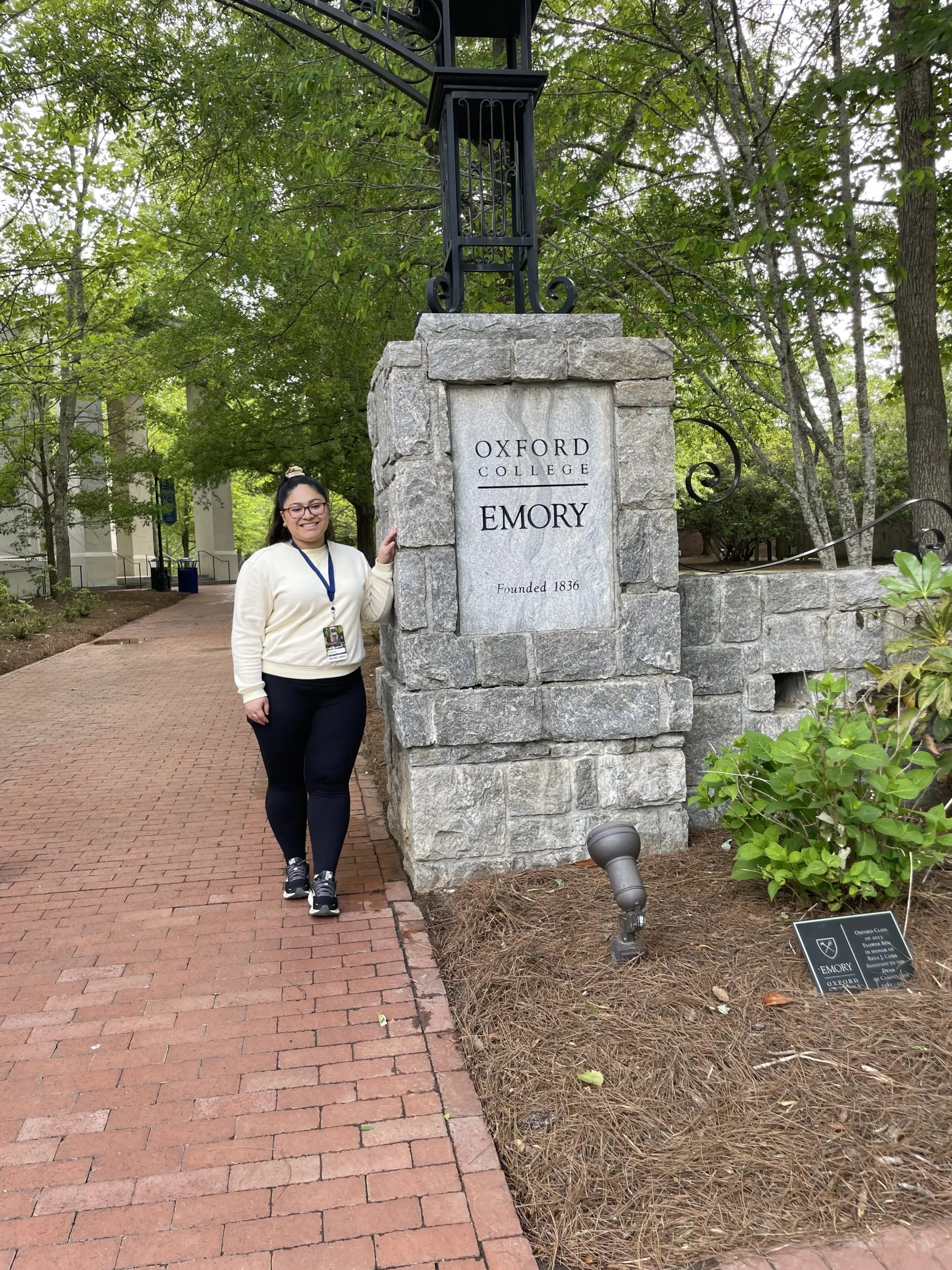 Affirmative action: Young Latino woman with long dark hair in black pants and white sweatshirt stands on red brick path next to stone pillar with Emory school sign with large green trees in the background.