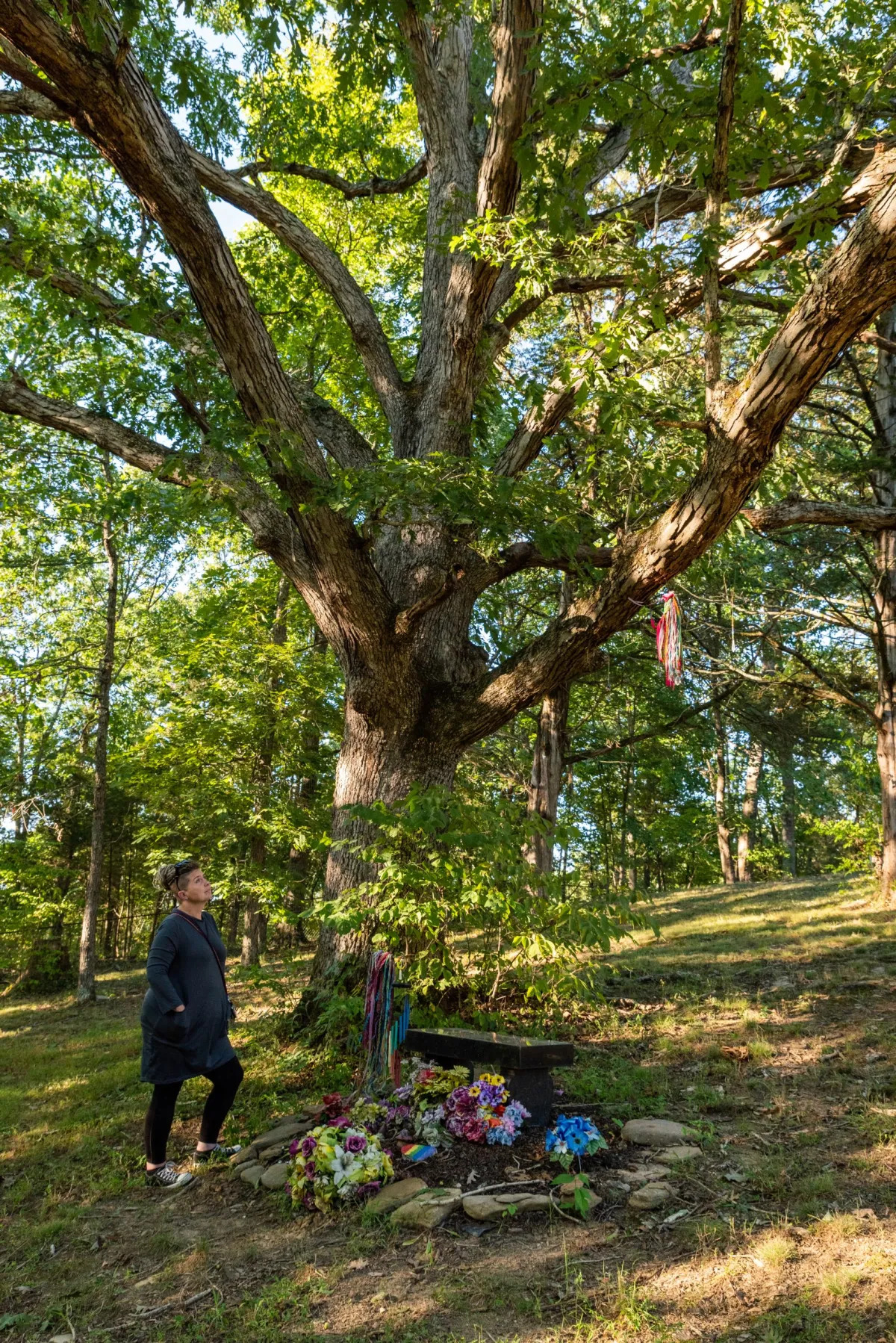 LGBTQ+ Clubs Kentucky: Woman standing next to planted flower bed and bench at memorial site on a hillside under a huge tree.
