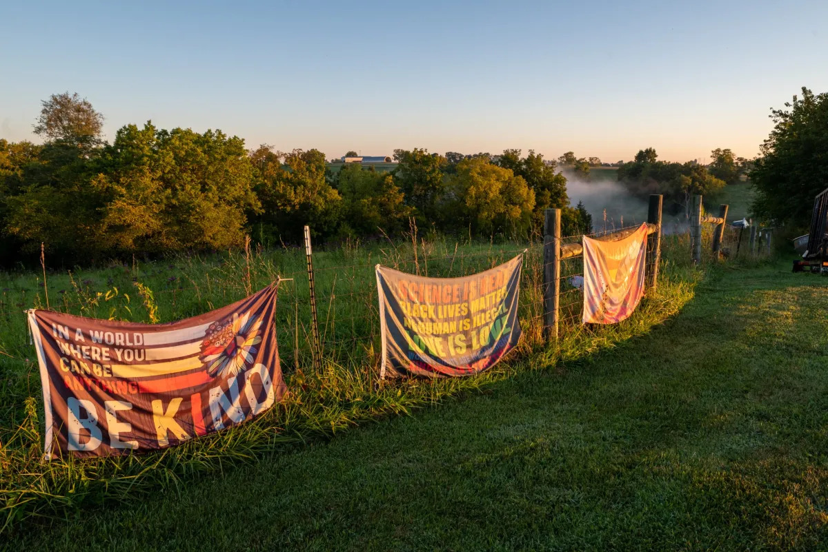 LGBTQ+ Clubs Kentucky: Wire fence in green pasture with line of big trees an lake in the background, with three large flags hanging on it.