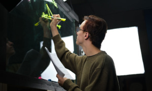 AI & education: Man with brown hair and black glasses in brown long-sleeved top stands with back to camera writing with yellow highlighter on glass blackboard, holding several papers in other hand.