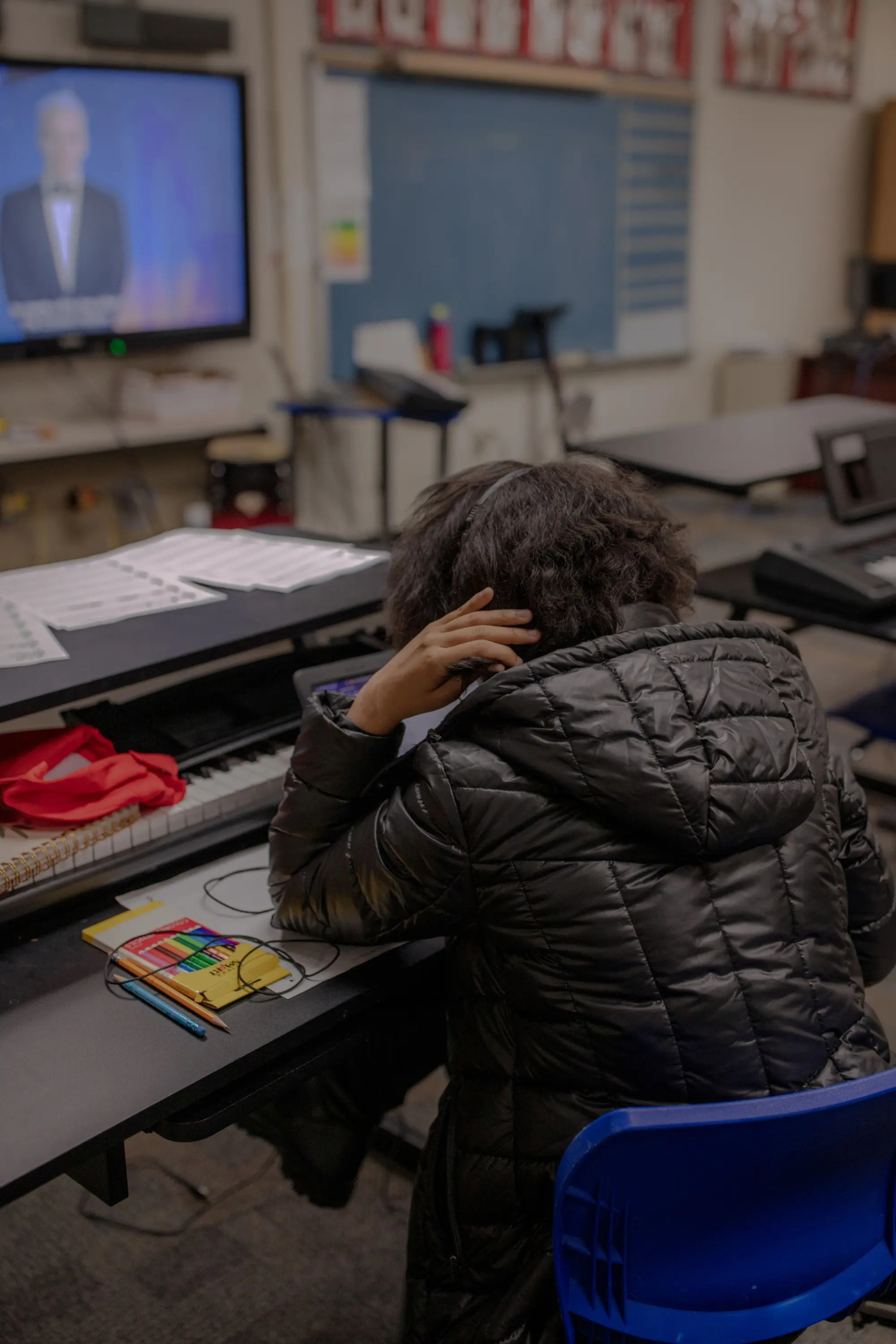 Foster teen mental health: Person with dark hair in quilted, black winter jacket sits at school desk with elbow on table and hand twirling hair at neck