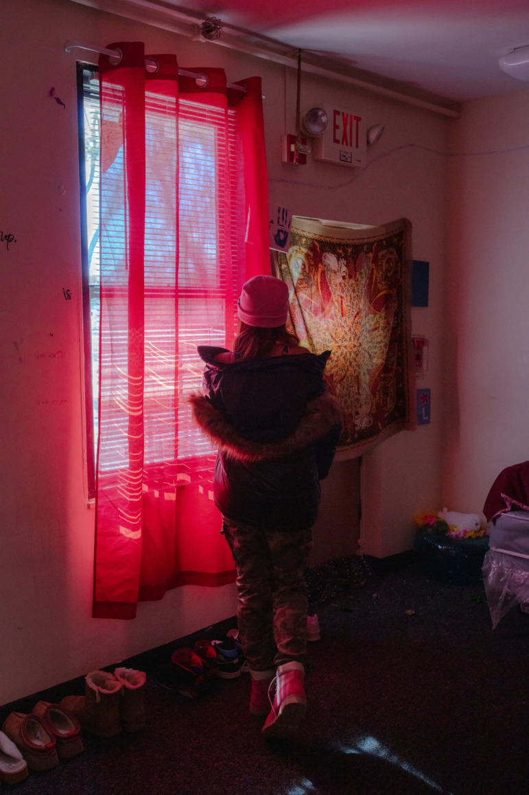 Foster teen mental health: Person in winter jacket and hat stands in dimly lit room, their back to camera, looking out of large paned window with sheer red drapes.