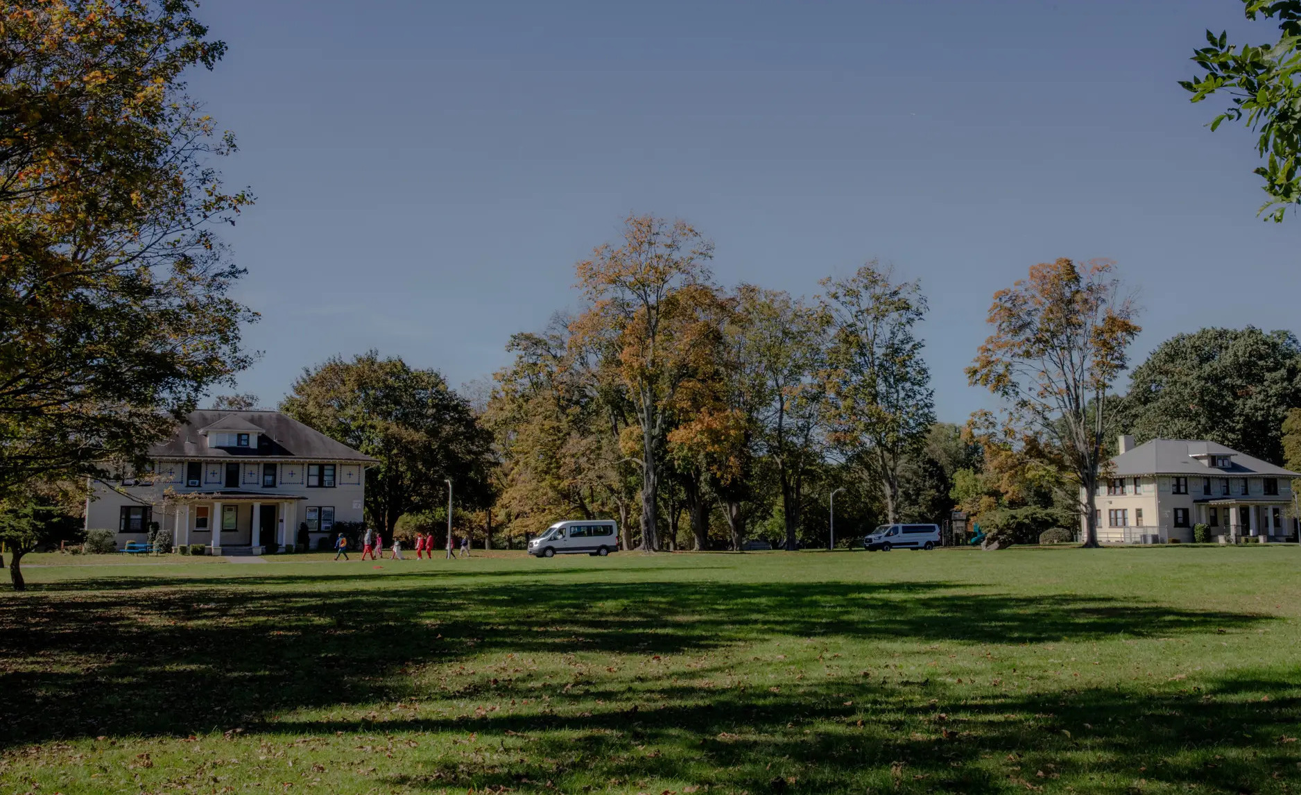 Foster teen mental health: Two traditional style, beige, 2-story buildings sit far back behind a huge lawn surrounded by tall, leady trees under clear blue sky.