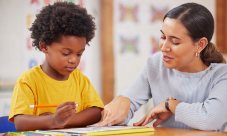 Reading tutoring: Woman with dark hair next to young Black elementary student in bright yellow t-shirt both looking at paper on desk