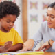 Reading tutoring: Woman with dark hair next to young Black elementary student in bright yellow t-shirt both looking at paper on desk