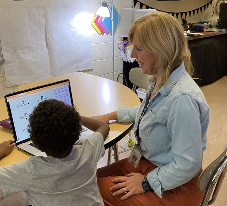 Reading tutoring: Woman with blonde hair sits next to young elementary student in white shirt both looking at laptop screen on desk