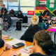 AI teacher evaluation: Classroom with several ethnically diverse students seated at desks and a female teacher facing the class standing in front of windows and colorful posters