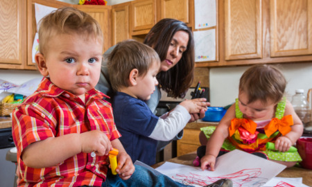 Childcare Utah: Woman caring for three toddlers sitting on kitchen island counter doing an art project in kitchen and baby in red plaid shirt with guilty face in foreground looks at camera