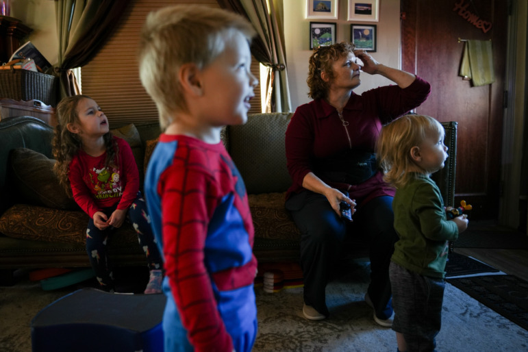 Utah childcare: Middle-aged woman with short red hair sits on a couch with one toddler and two other toddlers standing nearby watching TV