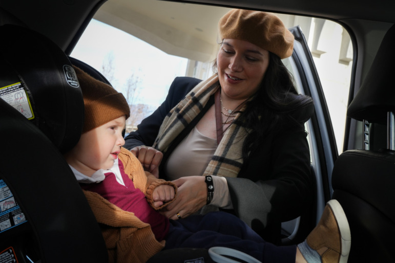 Utah childcare: Woman with long dark hair in tan beret and dark winter coat buckles a small toddler into a car sear.