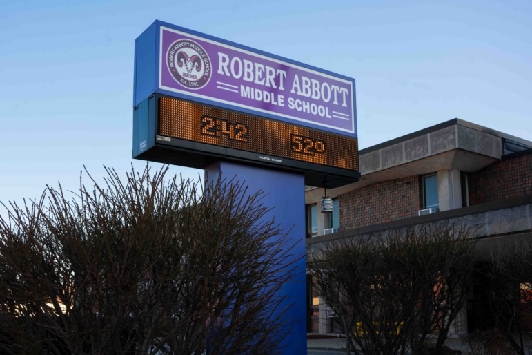 Brown & Black girls support Working on Womanhood: Large, tall, blue Robert Abbott Middle School school sign with LED text showing rime and temperature, in front of red brick building,