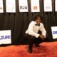 Atlanta student homelessness: Young Black man with black dreads in formal wear with black bow tie crouches on orange carpet in front of black curtain with several hanging paper signs.