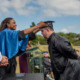 College access mentally disabled: Man with dark hair and beard in black college graduation gown leans forward while woman on bight blue cape places black college graduation cap on his head om an outdoor ceremony with trees on horizon