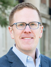 k-12 enrollment decline: Headshot of man with light brown hair and blue-framed glasses wearing blue suit smiles into camera