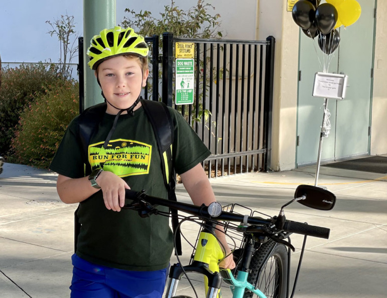 k-12 enrollment decline: Young boy os black r-shirt with bright yellow logo wearing neon yellow bike helme stands next to bike