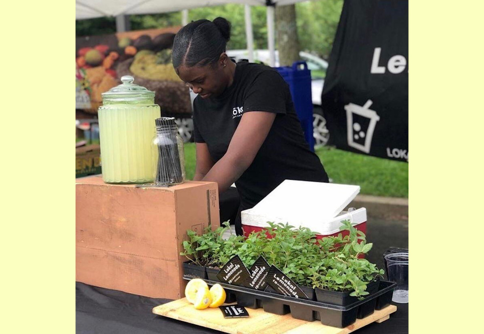 Youth Farmers Market: Black teen with black hair pulled back in navy t-shirt stands at table in farmers market with a box of fresh, leafy greens and glass pitcher of lemonade on display