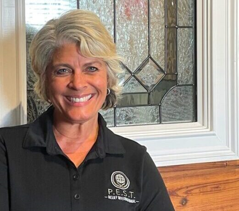 Everyday Heroes: Headshot Andrea Hancock middle-aged woman with short silver hair in black shirt with P.E.S.T. round logo in white, in front of leaded glass window of red brick building
