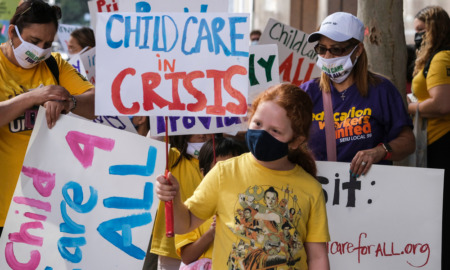 Childcare providers organizing: Protesrers with signs reading "Childcare Crisis" and "Childcare 4 All" gathered together outside on a sunny day