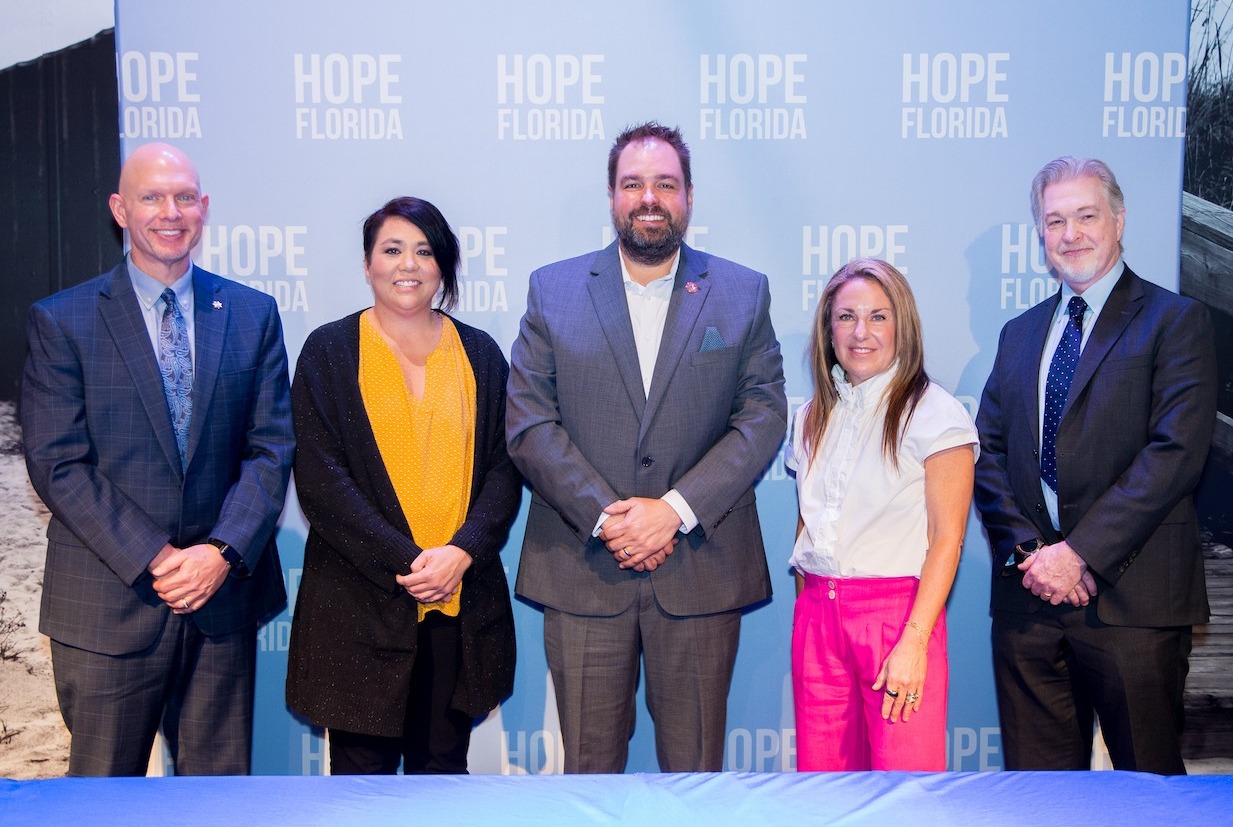 Mentoring month: 3 men and 2 women in business cloths stand in front of floor to ceiling light blue screen with white Hope Florida logo repeat printed across several rows