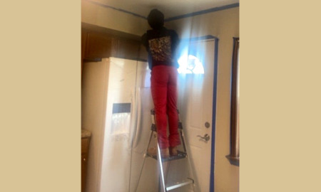 Trauma-informed hubs: Teen standing in ladder next to kitchen back door with back to camera folding tool and doing repairs to ceiling above refrigerator