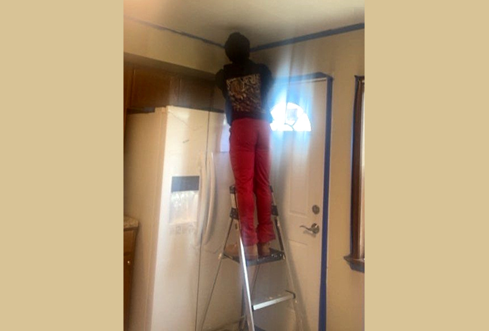 Trauma-informed hubs: Teen standing in ladder next to kitchen back door with back to camera folding tool and doing repairs to ceiling above refrigerator