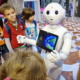 Robots in schools: White, child-sized, humanoid robot with computer monitor embedded in chest showing picture of earth, stands with hands outreached to group of elementary students surrounding it