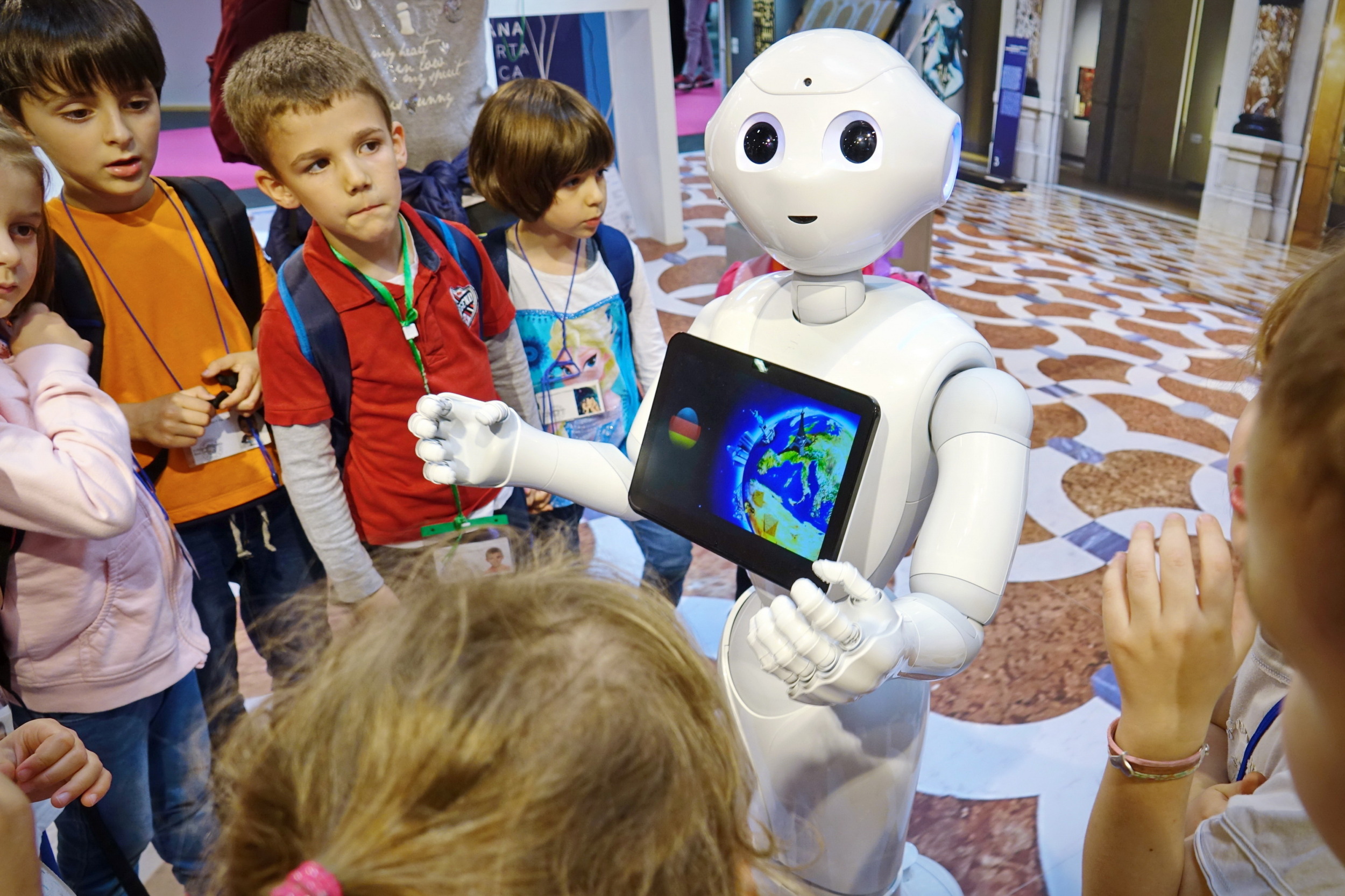 Robots in schools: White, child-sized, humanoid robot with computer monitor embedded in chest showing picture of earth, stands with hands outreached to group of elementary students surrounding it