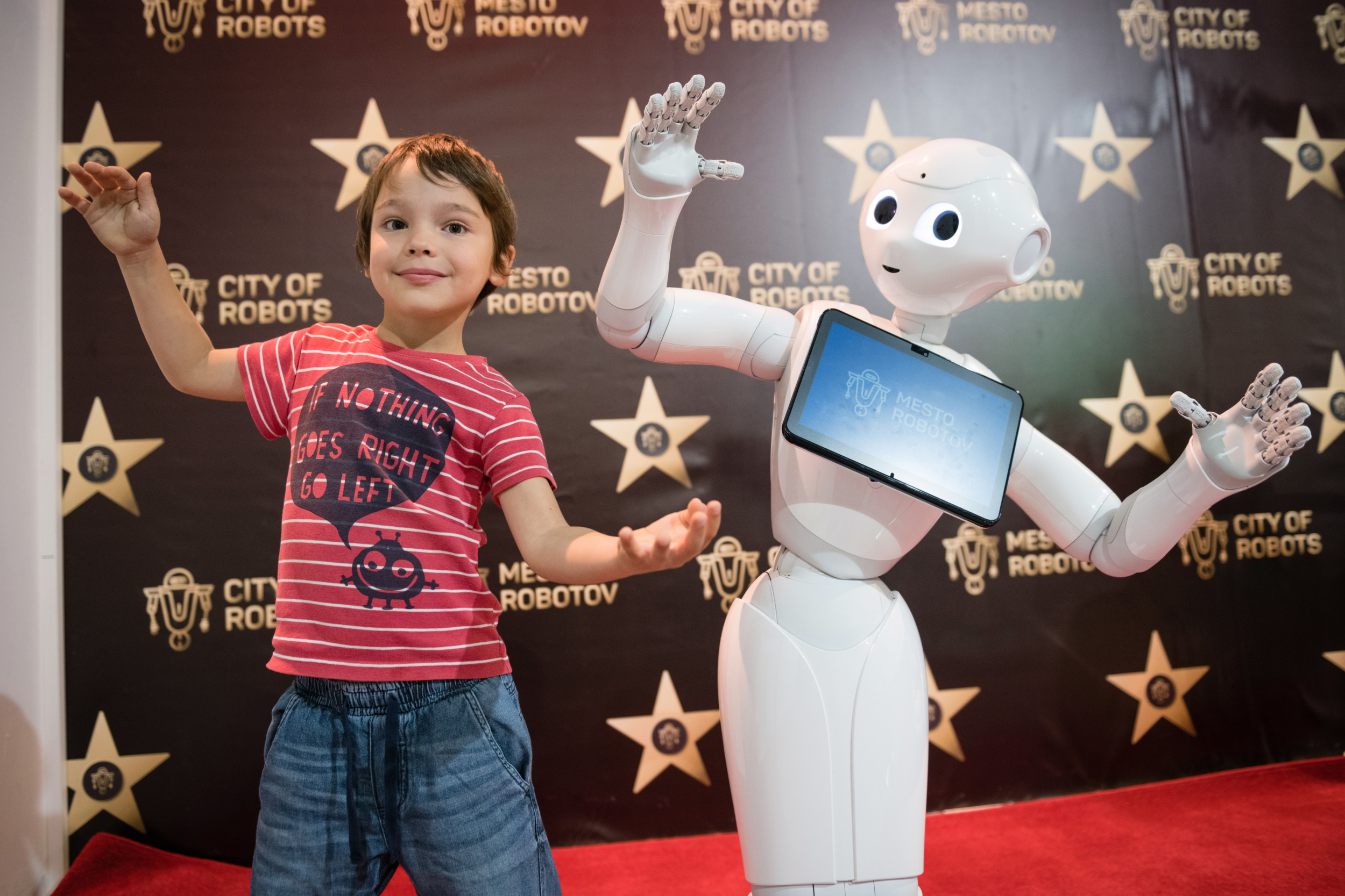 AI robots in school: Young boy in jeans and red t-shirt stands with arms outstretched to sides and leaning to his left, while white, humanoid, child-sized robot to child's left mimics the pose perfectly.
