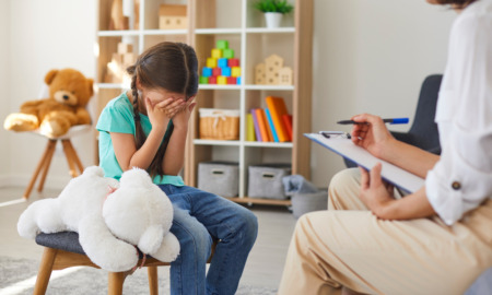 Sexual/child abuse recovery, child abuse professionals: Young girl sits on stool next to large, white, stuffed animal, hed down with hands covering her face across from woman with clipboard sitting on couch