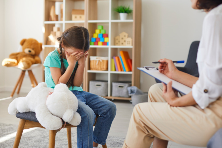 Sexual abuse recovery: Young girl sits on stool next to large, white, stuffed animal, head down with hands covering her face across from woman with clipboard sitting on couch
