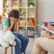 Sexual/child abuse recovery, child abuse professionals: Young girl sits on stool next to large, white, stuffed animal, hed down with hands covering her face across from woman with clipboard sitting on couch