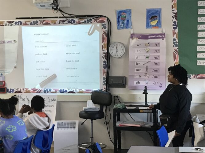 Literacy charter school: Teacher sits at front of classroom at her desk with elementary students sitting at school desks looking at white board