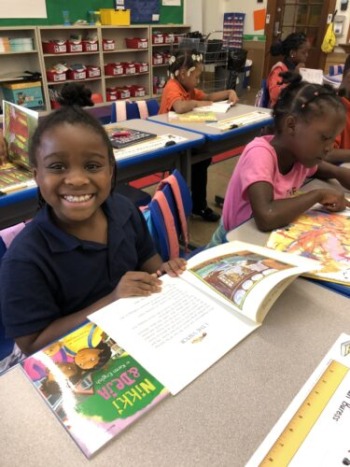 Literacy charter school: Smiling elementary student sits at school desk with large illustrated book open on desk