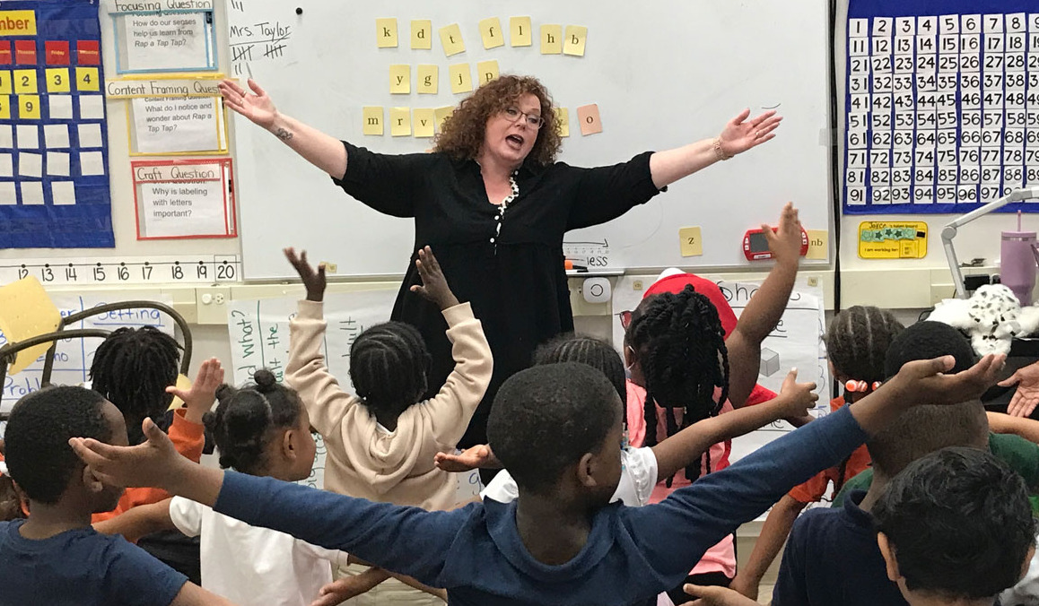Literacy charter school: Teacher stands with arms outstretched at head of class with several seated elementary students with arms outstretched