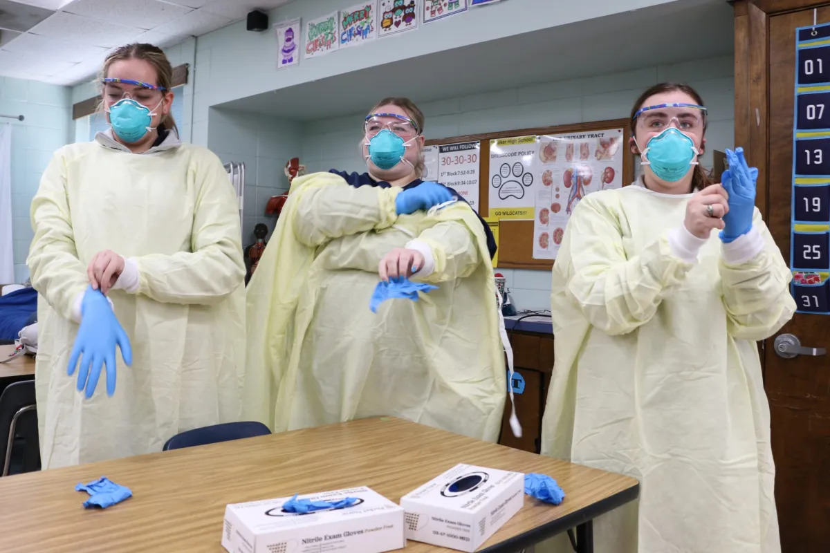 High school career and tech education: Three students in a high school classroom in yellow medical cover gowns, face masks and protective eye gear, stand at table pulling on blue latex gloves