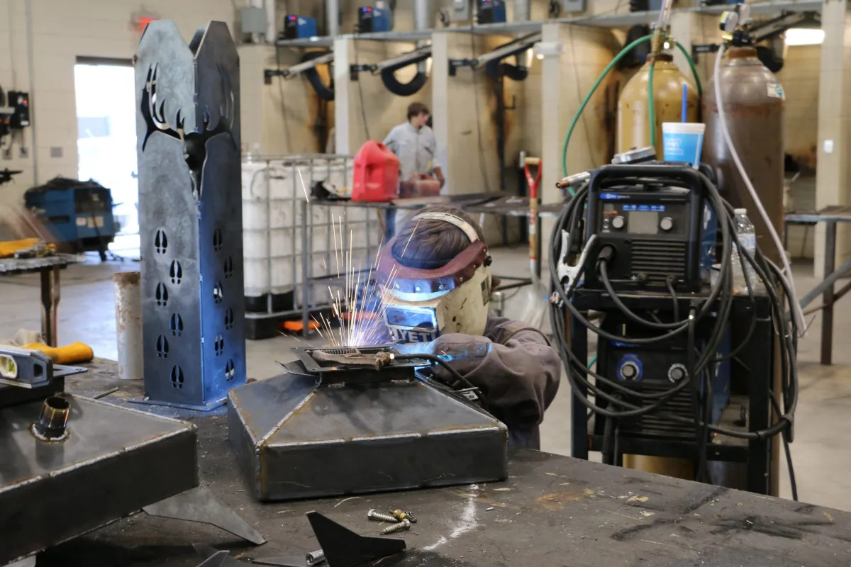 High school career and tech education: Student in protective head /eye gear works with welding equipment on a metal piece on a work table in a welding workroom full of equipment
